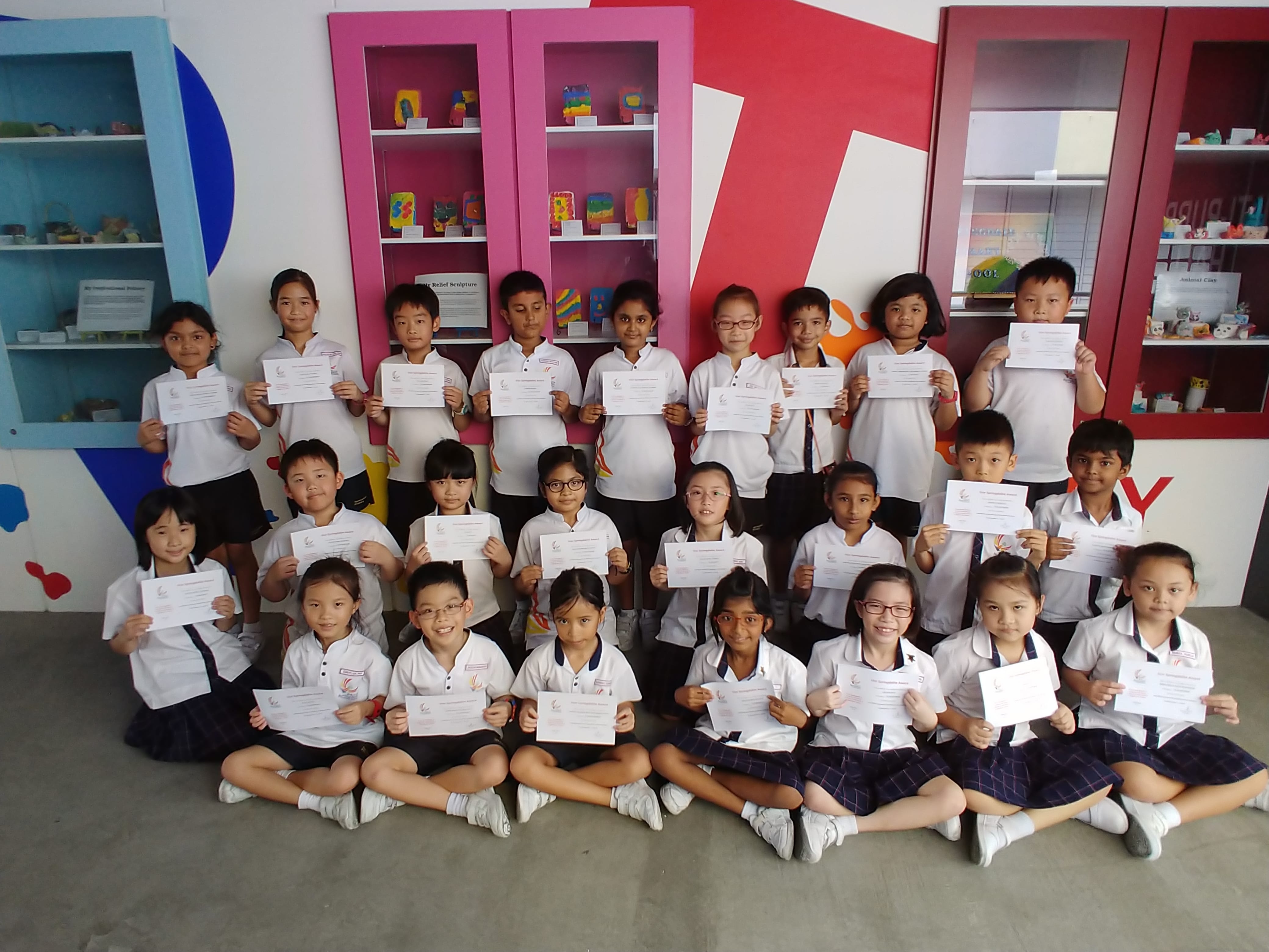 Group of young students in white uniforms holding certificates, posed in three rows in front of colorful display shelves.